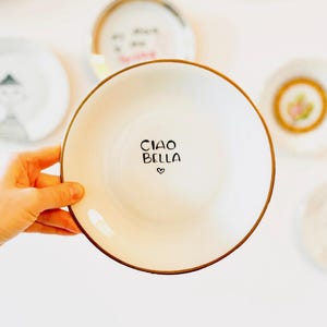 Könnte beinhalten: Ein weißer Keramikteller mit goldfarbenem Rand, auf dem der Text "Ciao Bella" in schwarzer Schrift mit einem kleinen Herzsymbol steht.