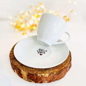 May include: A white ceramic teacup and saucer set. The saucer features the text "DU WIRST OPA" with a red heart. The set rests on a rustic wooden coaster, with a blurred background of golden lights.