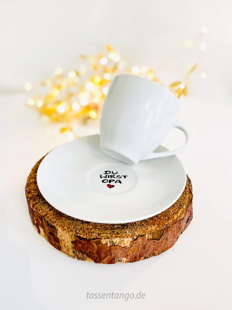 May include: A white ceramic teacup and saucer set. The saucer features the text "DU WIRST OPA" with a red heart. The set rests on a rustic wooden coaster, with a blurred background of golden lights.