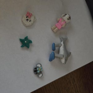 May include: A collection of five small decorative charms including a white bear face with a pink bow, a small figure in a pink dress, a teal star with a center stud, a gray shark with blue fins, and a gray koala face with a light blue flower.