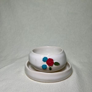 Pode incluir: Um vaso de cerâmica branco com um pires a condizer. O vaso apresenta um design floral em relevo com uma rosa vermelha, duas flores azuis e folhas verdes. O vaso é redondo e tem um acabamento liso e brilhante. Ideal para plantas pequenas.