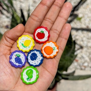 Pode incluir: Seis pequenos pingentes redondos com bordas recortadas, apresentando desenhos de frutas pintados à mão sobre fundos brancos com pontos coloridos. A coleção inclui uma maçã vermelha, um limão amarelo, uvas roxas, um mirtilo azul, uma laranja e uma pera verde.