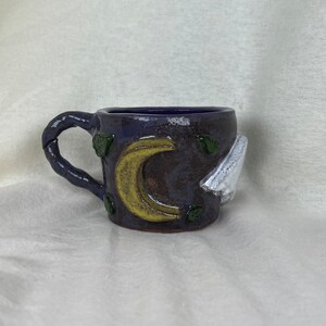 Pode incluir: Uma caneca de cerâmica artesanal com exterior azul escuro, apresentando um design de lua crescente amarela e nuvem branca. A caneca tem uma alça texturizada e detalhes de folhas verdes. Uma peça única para desfrutar de bebidas.