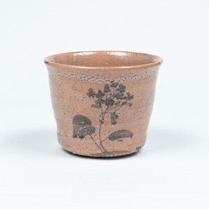 May include: A small, brown ceramic cup with a black floral design. The cup has a textured surface and a slightly flared rim.