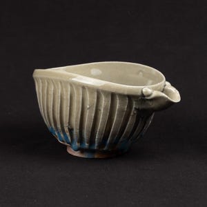 May include: A ceramic bowl with a spout, showing a textured, ribbed design on the outside. The bowl is mainly a light beige colour, with blue and brown accents at the base. The spout is small and rounded.