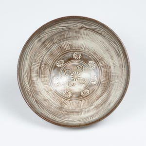 May include: A ceramic bowl with a detailed, textured design. The bowl features concentric circles and floral patterns in a light brown and white color scheme. The rim has a darker brown hue.