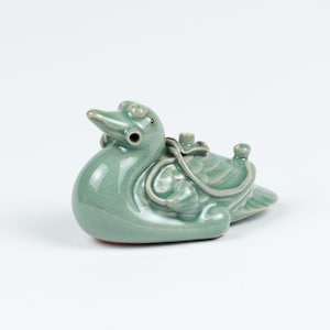 Puede incluir: Figura de pato de cerámica esmaltada celadón. El pato tiene un diseño detallado con un cuello curvo, un pico pequeño y un elemento decorativo en la espalda. La figura es de color verde claro.