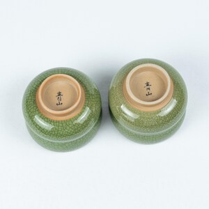 May include: Two green ceramic bowls with a crackled glaze. The bowls have a brown interior with Japanese characters written in black.