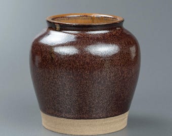 May include: A handcrafted, brown ceramic jar with a speckled texture and a glossy finish. The jar has a wide, rounded body and a slightly flared rim. The base is a lighter, unglazed tan color. This decorative piece is suitable for home decor.