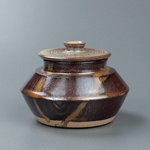 Peut inclure: Un pot en céramique fait à la main avec un couvercle. Le pot est de couleur brun foncé avec des stries marron clair et beige. Le couvercle présente des cercles concentriques et un petit bouton. Le pot repose sur une base beige clair.