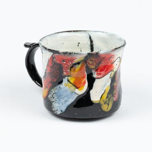 Könnte beinhalten: Eine schwarze Keramiktasse mit weißem Innenraum und schwarzem Henkel. Die Tasse ist mit abstrakten Formen in Rot, Orange, Gelb, Blau und Weiß verziert.