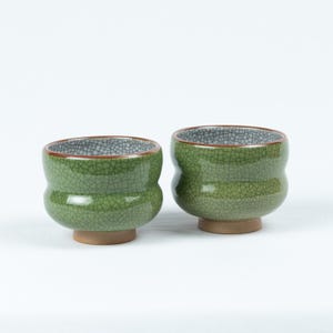 Op de afbeelding: Twee groene keramische theekopjes met een gekrakelde glazuur. De kopjes hebben een afgeronde vorm en een klein voetje.