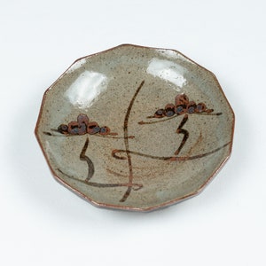 May include: A small, light brown ceramic dish with a scalloped edge. The dish has a brown and black design of stylised branches and clusters of brown dots.