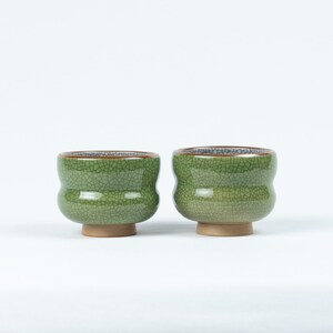 May include: Two green ceramic teacups with a crackled glaze and brown rims. The cups have a rounded shape and a small foot.