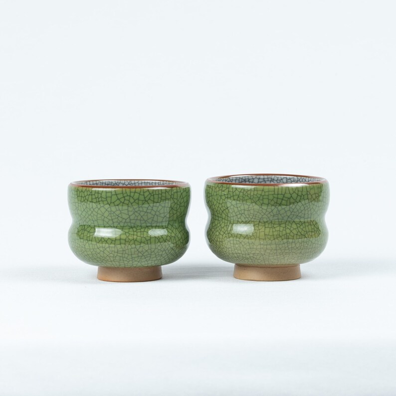 May include: Two green ceramic teacups with a crackled glaze and brown rims. The cups have a rounded shape and a small foot.