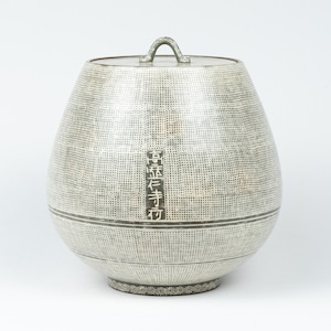 Op de afbeelding: Een zilverkleurige container met deksel, een getextureerd oppervlak en een klein handvat. De container heeft een afgeronde vorm met een decoratief bloemmotief rond de basis. Verticale Japanse tekst is zichtbaar.