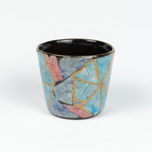 May include: A small ceramic cup with a mosaic design. The cup has a black interior and a textured exterior with blue, pink, and grey segments, outlined with gold lines. A decorative piece for the home.