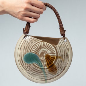 May include: A ceramic wall vase with a brown leather handle. The vase is decorated with a brown, white, and blue swirl pattern.