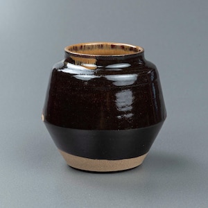 Può includere: Un vaso in ceramica con un corpo largo e angolare e un collo corto e stretto. La parte superiore presenta una smaltatura lucida marrone scuro che transita verso una base non smaltata color beige. Il bordo mostra un sottile effetto a goccia.