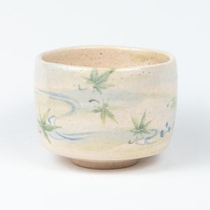 May include: A cream-coloured ceramic tea bowl. It features a hand-painted design of green maple leaves and blue water patterns. The bowl has a slightly flared rim and a small, unglazed foot. The bowl is approximately 8 cm in diameter.