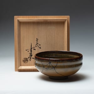 May include: A ceramic tea bowl with a brown and tan glaze, featuring visible kintsugi repairs. The bowl sits in front of a wooden box with Japanese calligraphy. The bowl's diameter is approximately 5 inches.