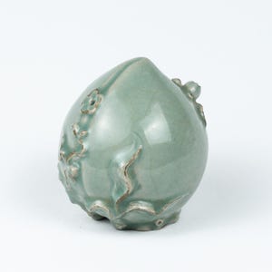 May include: A celadon-glazed ceramic peach-shaped decorative object. The object has a smooth, rounded form with embossed floral and leaf details. The surface has a crackle effect, and the color is a soft, muted green.