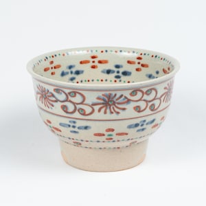 May include: A cream-coloured ceramic bowl with a wider top and a base. It features red, blue, and green floral and geometric patterns. The rim has small dots. The bowl's design is unique. The bowl's measurements are in centimetres.