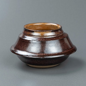 May include: A low-profile ceramic bowl with a wide, flared midsection and a narrow, circular opening. The exterior features a deep, glossy dark brown glaze with subtle black speckling, while the interior rim displays a lighter tan finish.