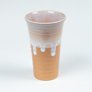 May include: A tall, handmade ceramic tumbler with a textured, ridged base. The cup transitions from a light brown to a beige color, with a white glaze dripping down the sides. The rim is a light beige color.