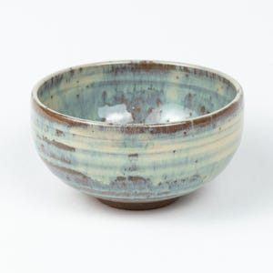 May include: A handcrafted ceramic bowl with a distinctive glaze. The bowl has a light blue and cream colour scheme with brown accents around the rim and base. The bowl is round and suitable for serving food.
