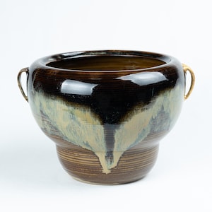 Peut inclure: Un grand pot en céramique brun avec deux poignées dorées. Le pot a un glaçage unique qui crée un effet marbré avec des nuances de brun, de noir et de blanc.