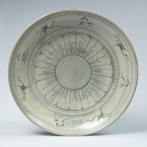 May include: A large, white ceramic plate with a green glaze. The plate is decorated with a central sunburst design and a border of stylized cranes.
