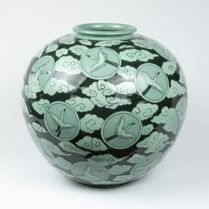 May include: A round, celadon-colored ceramic vase with a dark green background. The vase features a repeating pattern of white cranes in flight, set within circular frames, and surrounded by stylized clouds. The vase has a wide body and a narrow neck.