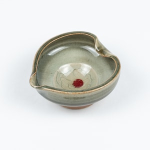 May include: A small, heart-shaped ceramic bowl with a green glaze and a red dot in the centre. The bowl has a slightly raised rim and a small spout on one side.