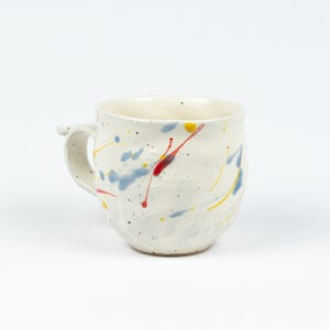 Könnte beinhalten: Handgefertigte Keramik-Tasse mit weißem Grund und farbenfrohem Farbspritzer-Design. Die Tasse hat blaue, rote und gelbe Farbspritzer. Sie hat einen kleinen Henkel und eine strukturierte Oberfläche. Geeignet für Kaffee, Tee oder andere Getränke.