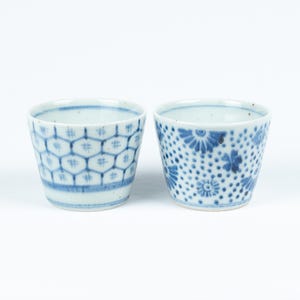 May include: Two small white ceramic cups with blue and white patterns. The cup on the left has a geometric pattern, while the cup on the right has a floral pattern.