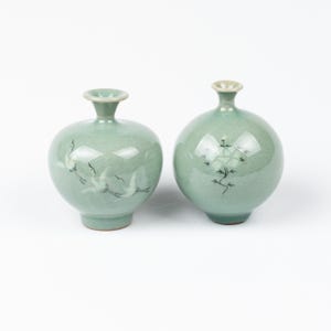 Puede incluir: Dos jarrones de cerámica color celadón con acabado brillante. Un jarrón presenta un diseño de grulla, el otro un motivo floral. Ambos tienen un cuerpo redondeado, un cuello estrecho y una pequeña abertura.