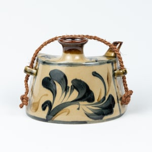 May include: A ceramic sake bottle with a beige base and dark blue floral design. The bottle has a brown rim and spout, with a brown rope handle. The bottle is a unique piece of pottery.