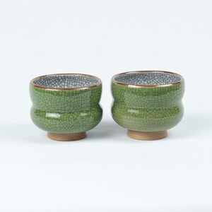 May include: Two green ceramic teacups with a crackled glaze. The cups have a rounded shape and a brown rim.