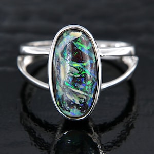 Könnte beinhalten: Ein Silberring mit einem ovalen Opal-Edelstein. Der Opal zeigt ein lebendiges Farbspiel mit grünen, blauen und roten Reflexen auf dunklem Hintergrund. Das Band des Rings hat ein geteiltes Design, das dem Schmuck eine moderne Note verleiht.