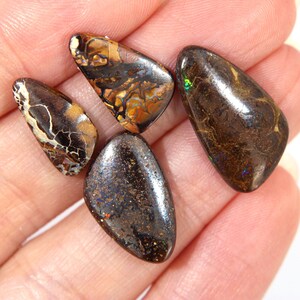 Pode incluir: Quatro opalas boulder polidas com formas irregulares semelhantes a gotas. As pedras apresentam uma matriz de pedra de ferro castanho-escura com padrões rodopiantes de cores iridescentes, incluindo reflexos verdes, azuis e dourados.