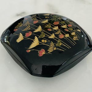 May include: A black, lacquered box with a floral design. The box has a rounded, geometric shape and is decorated with gold, red, and green flowers and leaves. The box is sitting on a white marble surface.