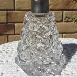 May include: A clear glass bottle with a faceted design and a silver metal top. The bottle has a square base and a tall, narrow neck.