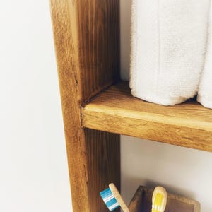May include: A wooden shelf unit with two shelves. The top shelf has two white towels folded on it. The bottom shelf has two toothbrushes.