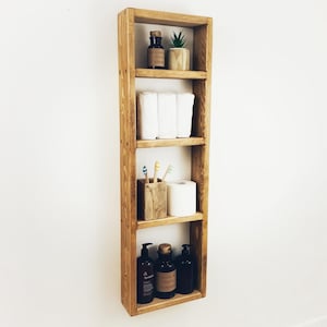 May include: A wooden shelf unit with four shelves, featuring a small potted plant, toilet paper, toiletries, and a toothbrush holder. The shelf is mounted on a white wall.