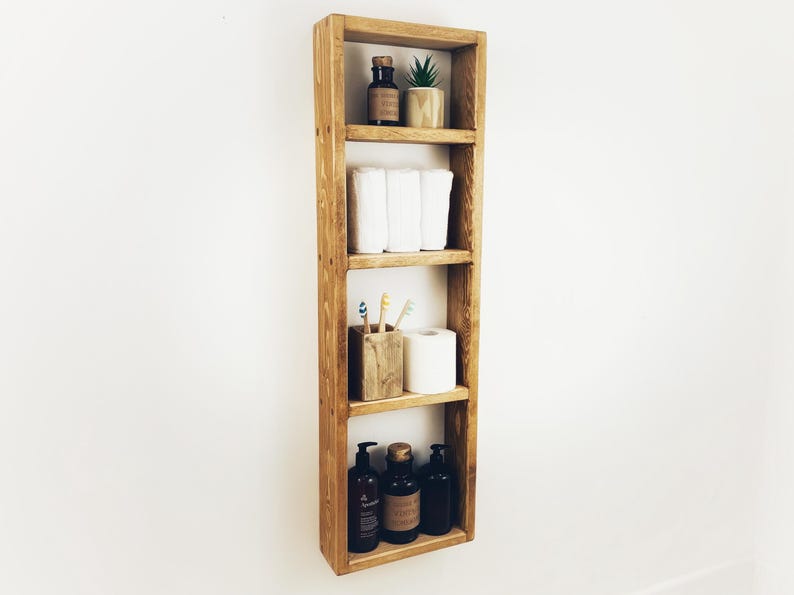 May include: A wooden shelf unit with four shelves, featuring a small potted plant, toilet paper, toiletries, and a toothbrush holder. The shelf is mounted on a white wall.