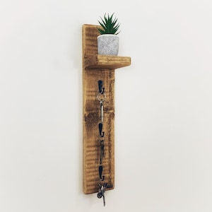 May include: A rustic wooden wall mounted shelf with a small potted succulent plant on the top shelf. The shelf has four hooks on the front for hanging keys or other items.