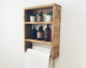 May include: A rustic wooden shelf with three shelves and a towel bar. The shelf is filled with small bottles, plants, and a towel.