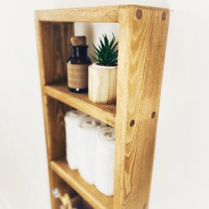 May include: A wooden shelf unit with four shelves, featuring a small potted succulent plant, a glass bottle, rolled towels, and a roll of toilet paper. The shelf unit is stained in a light brown color and has a rustic appearance.