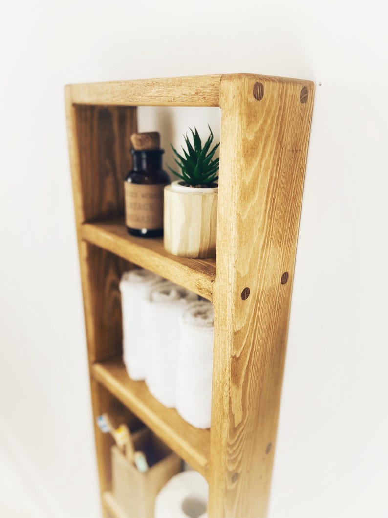 May include: A wooden shelf unit with four shelves, featuring a small potted succulent plant, a glass bottle, rolled towels, and a roll of toilet paper. The shelf unit is stained in a light brown color and has a rustic appearance.